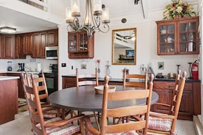 A dining area with a round table, wooden chairs, a chandelier, and a kitchen in the background. - Beech Mtn Chalet | Long-Range Views, Sleeps 9, AC (Beech Mountain)