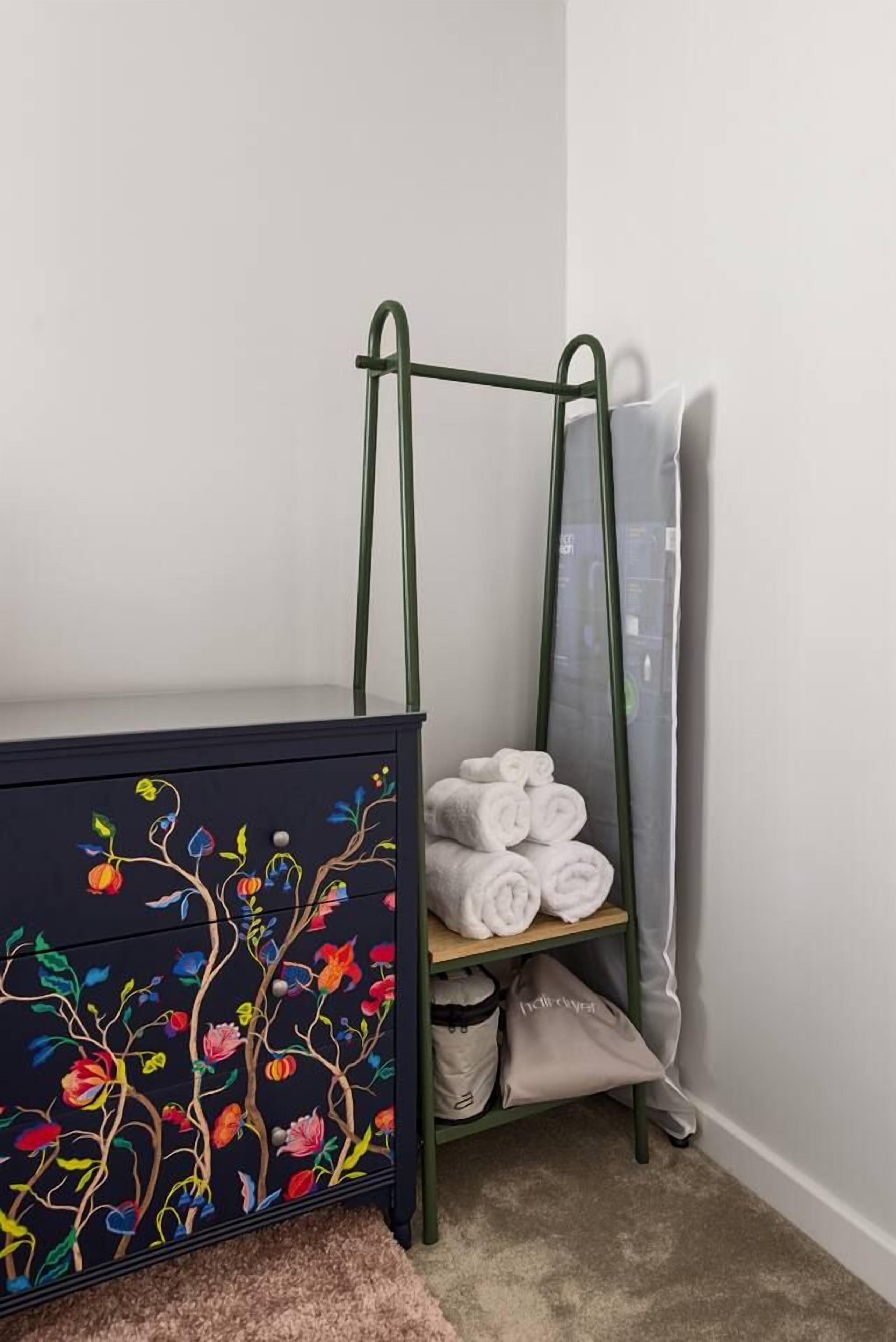 Een kamer met een donker kastje met bloemmotieven, een groen metalen rek met handdoeken, en een grijze gordijn.