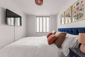 Un dormitorio con cama, televisión montada en la pared, ventana con persianas y cabecera azul.