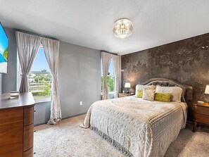 A bedroom with a large bed, two bedside tables with lamps, a dresser, and a view of greenery through the windows. - Snowbird Special: Mint 3BR Gated Townhome, Seasonal Stay Available (Stuart)