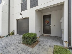 A modern building with a garage door, a front door with a flower wreath, and a small garden area with a bush. - Snowbird Special: Mint 3BR Gated Townhome, Seasonal Stay Available (Stuart)