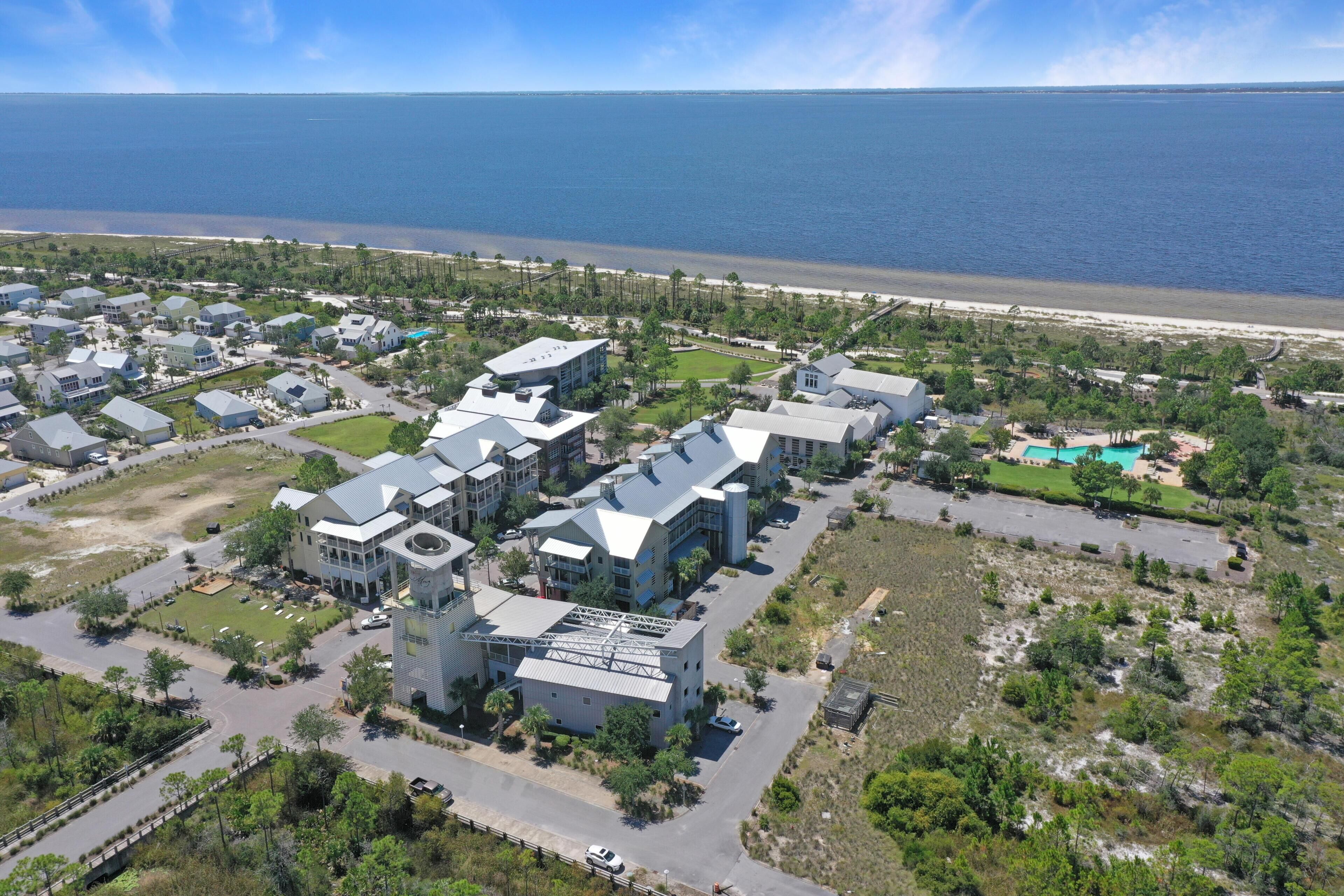 Vista aérea de una zona residencial costera con casas modernas, un edificio grande con una estructura de techo distintiva y una playa al fondo.
