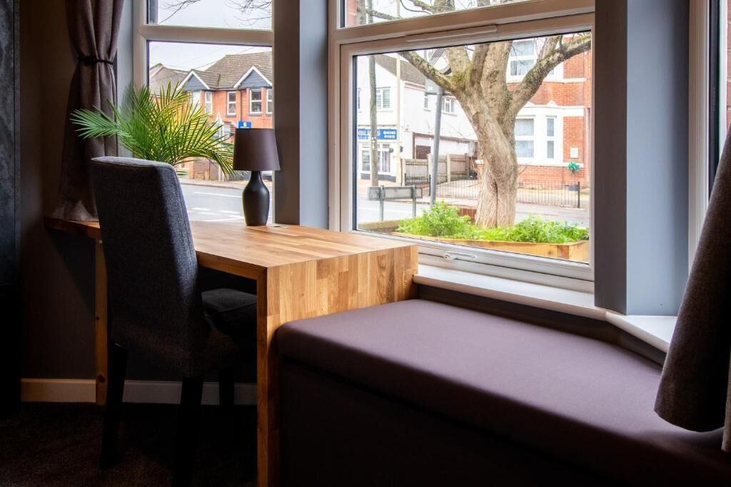 Une pièce avec un bureau en bois, une chaise, un banc, une lampe et une vue sur la rue et les bâtiments environnants.