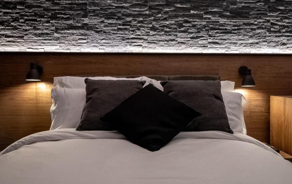 A bed with white and dark pillows, two black wall-mounted lamps, and a stone-textured wall above.