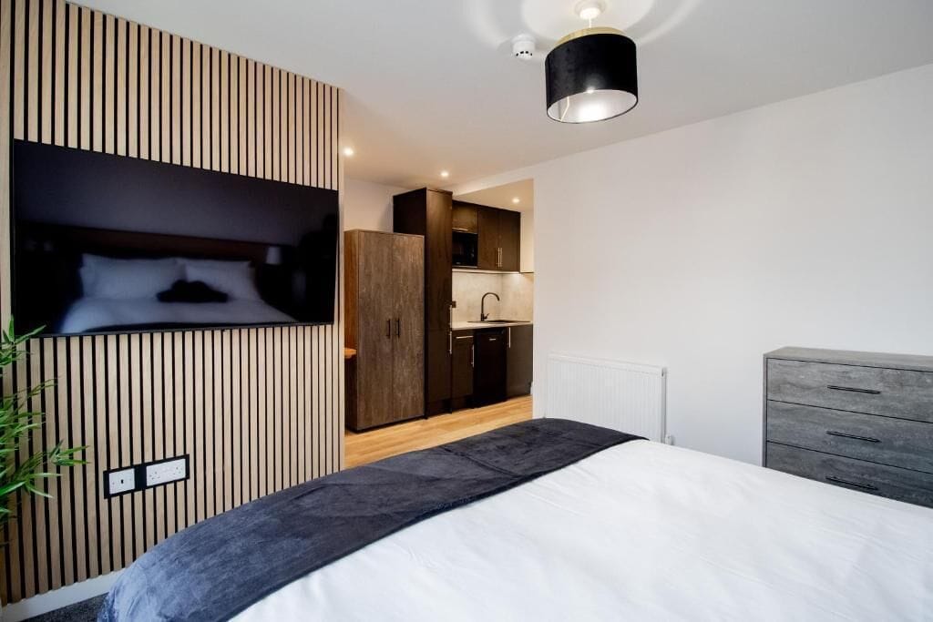 Une chambre moderne avec un lit, un téléviseur fixé au mur, qui est recouvert d’un panneau en bois, une commode grise et, au fond, un coin cuisine.