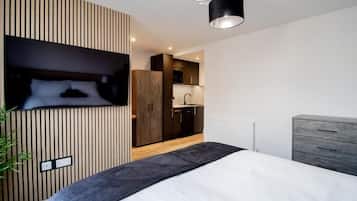 Une chambre moderne avec un lit, un téléviseur fixé au mur, qui est recouvert d’un panneau en bois, une commode grise et, au fond, un coin cuisine.