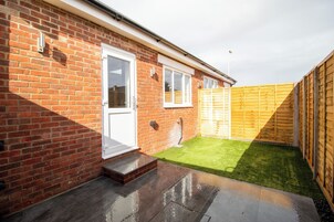 A brick house with a white door, a small front yard with artificial grass, and a wooden fence.