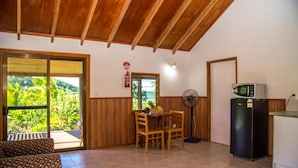 A room with a wooden ceiling, a dining table with chairs, a fan, a refrigerator, and a sliding glass door leading to a balcony with a view of greenery.