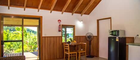 A room with a wooden ceiling, a dining table with chairs, a fan, a refrigerator, and a sliding glass door leading to a balcony with a view of greenery.