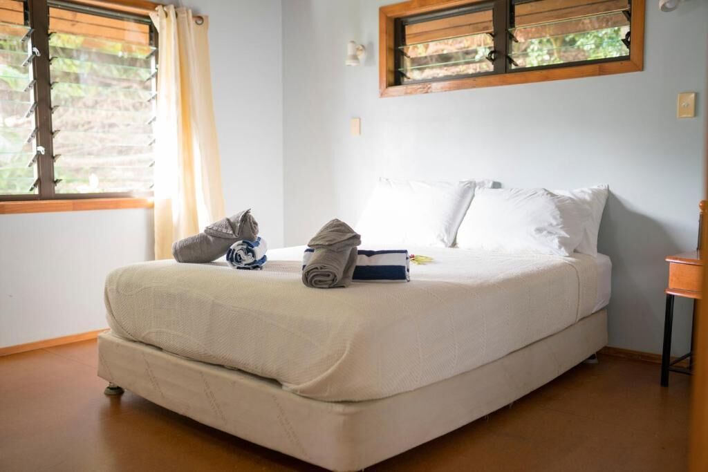 A single bed with a white mattress and pillows, a rolled towel, and a small bag on the bed. There is a window with blinds on the left side of the bed.