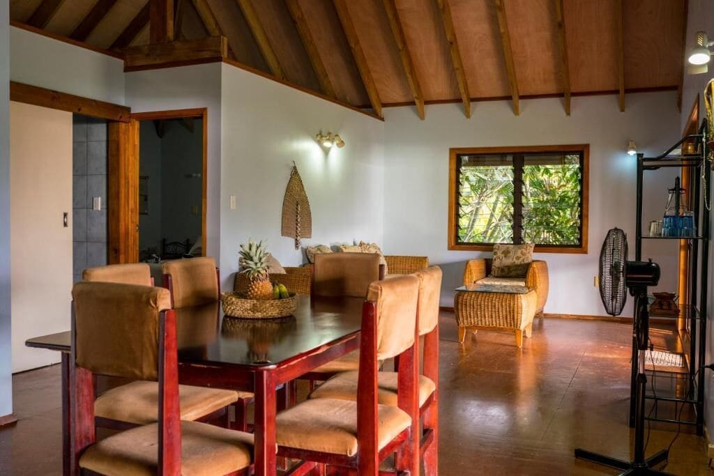 A dining area with a wooden table and chairs, a wicker sofa, and a window with a view of greenery.