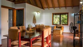 A dining area with a wooden table and chairs, a wicker sofa, and a window with a view of greenery.