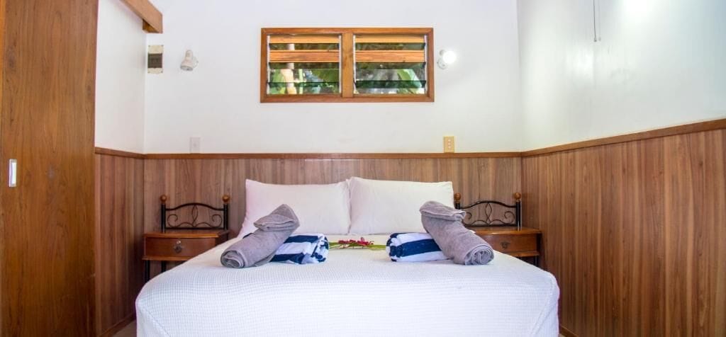 A hotel room with a wooden paneled wall, a bed with white linens, two bedside tables, and a window with wooden shutters.