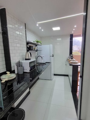 A modern kitchen with a black countertop, white appliances, and a view of the city through a window. - Loft Fátima (Teresópolis)
