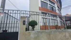 Un edificio a più piani con un ingresso chiuso da cancello, un piccolo albero davanti e un muro in cemento.