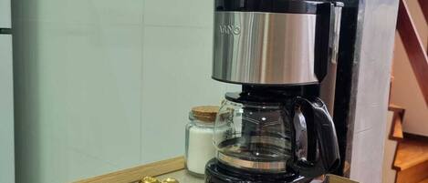 A coffee maker on a wooden tray with a marble countertop, a jar of sugar, and a pair of golden spoons.