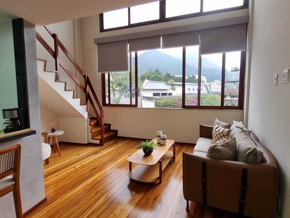 A spacious living area with a wooden floor, a staircase, a sofa, and a large window offering a view of buildings and mountains.