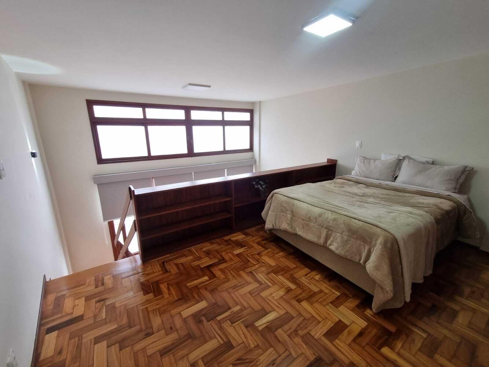 Ein Schlafzimmer mit Holzdielen-Parkett, einem Bett mit beiger Bettdecke, einem Fenster mit Rollläden und einer Heizung.