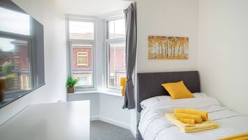 A hotel room with a bed, a desk, a window with a view of buildings, and a painting on the wall.