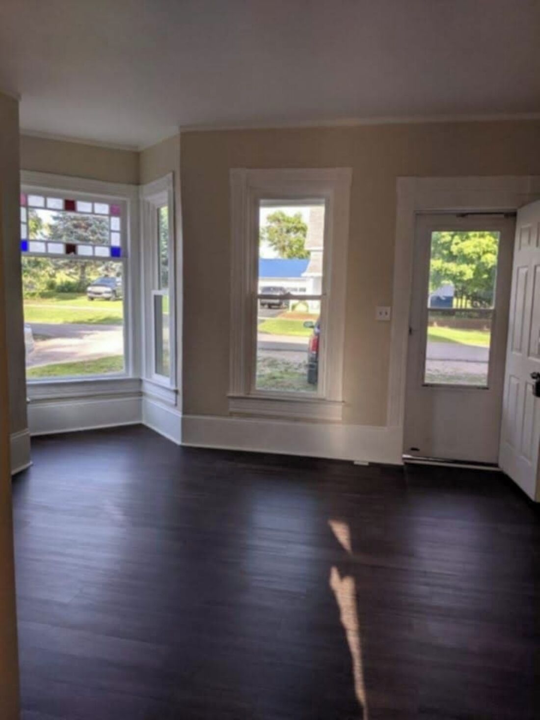 A room with dark wooden flooring, a white door, and a window with a view of a street and greenery.