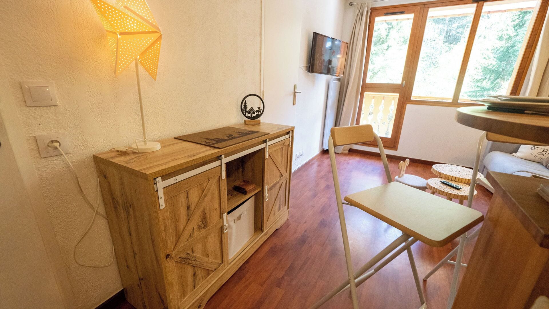 Habitación con escritorio de madera, una silla, una mesita con control remoto, televisor en la pared y ventana con cortinas.