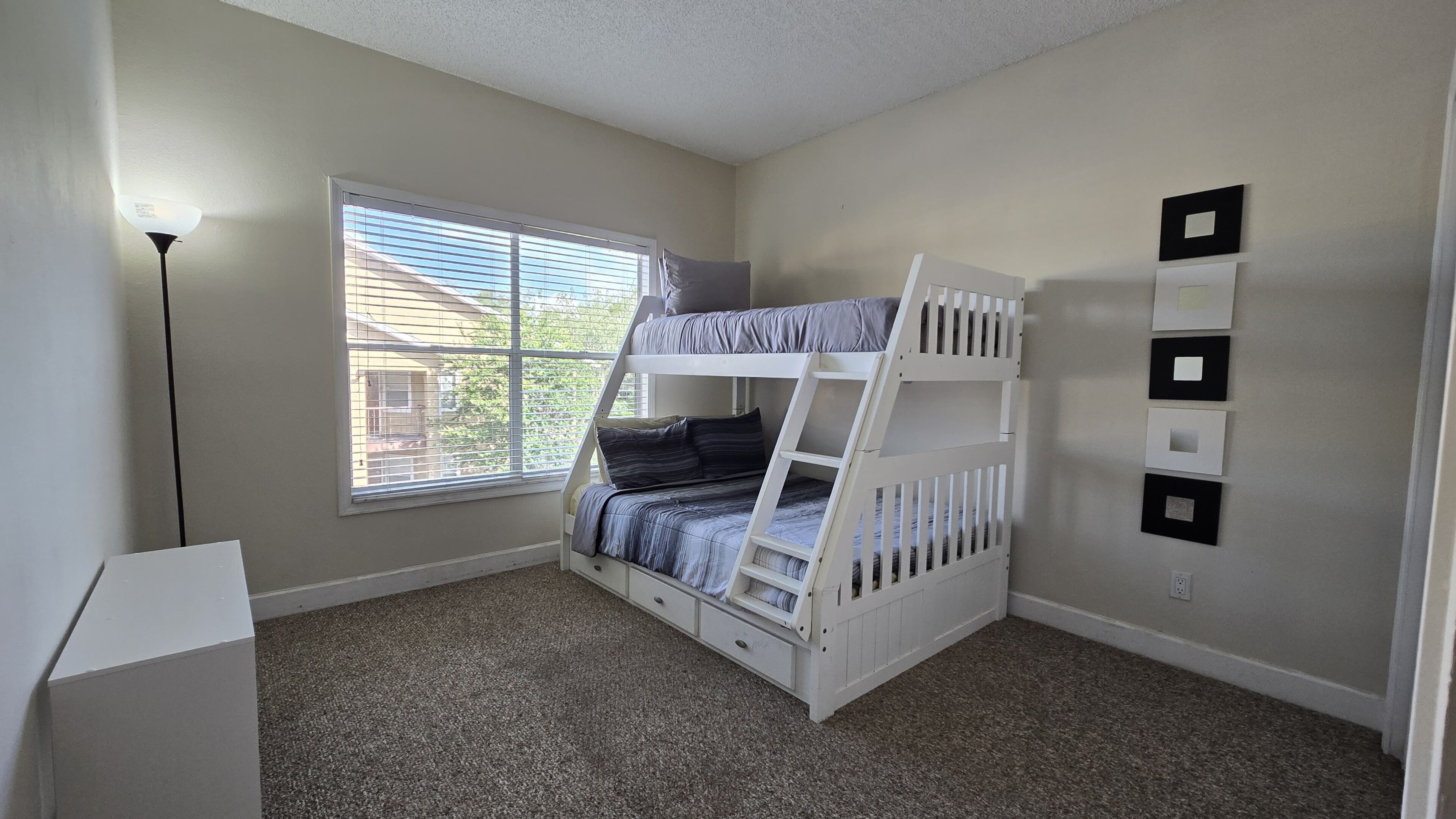 A room with a bunk bed, a window with blinds, a lamp, and a wall-mounted light switch.