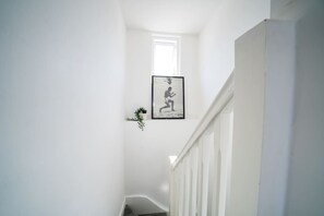 Un escalier étroit, avec sur le mur une photo encadrée représentant une personne accroupie, une petite plante en pot et une rambarde blanche.