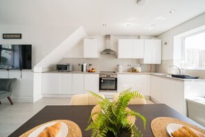 A modern kitchen with white cabinets, a microwave, and a stove. There is a dining table with a plant and bread. - Premium 3 Bed Cardiff City Sleeps 9 with Free Parking (Cardiff)
