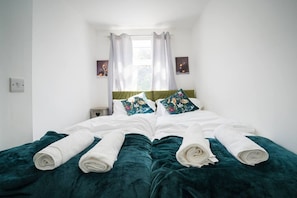 Ein Bett mit weißen Laken und türkisfarbenem Bettbezug, ein Nachttisch mit einem herzförmigen Gegenstand und ein Fenster mit weißen Vorhängen.
