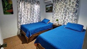 A room with two beds, each with blue bedding, white curtains with a leaf pattern, a painting on the wall, and an air conditioning unit. - Peace and Nature House. (La Fortuna)