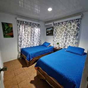 Quarto com duas camas, cada uma com roupa de cama azul, cortinas brancas com padrão de folhas, um quadro na parede e um aparelho de ar condicionado.