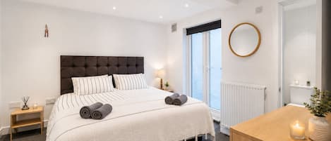 Une chambre moderne avec un grand lit, des tables de chevet, une commode en bois, un miroir rond et une fenêtre donnant sur un beau paysage.