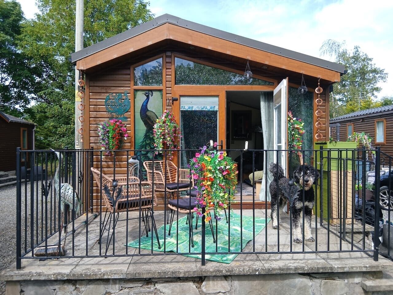 A wooden cabin with a glass door, a fenced patio with chairs, and a dog.