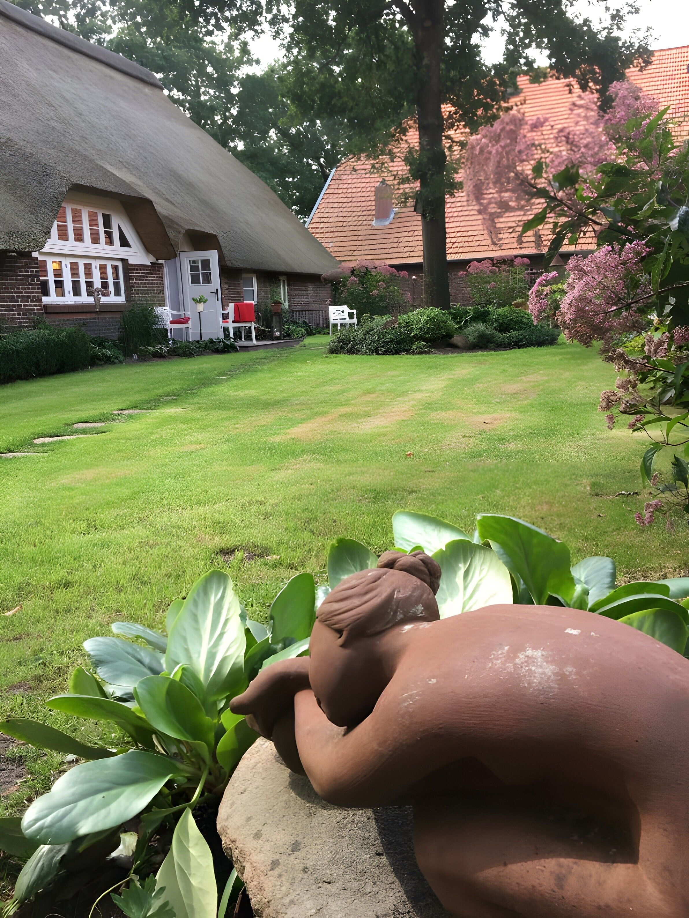 Ein Garten mit einem Haus mit Strohdach, einer Rasenfläche und einer Steinskulptur.