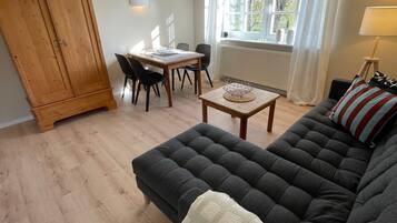 A living room with a dark grey sofa, a wooden dining table with chairs, a wooden wardrobe, and a window with curtains.