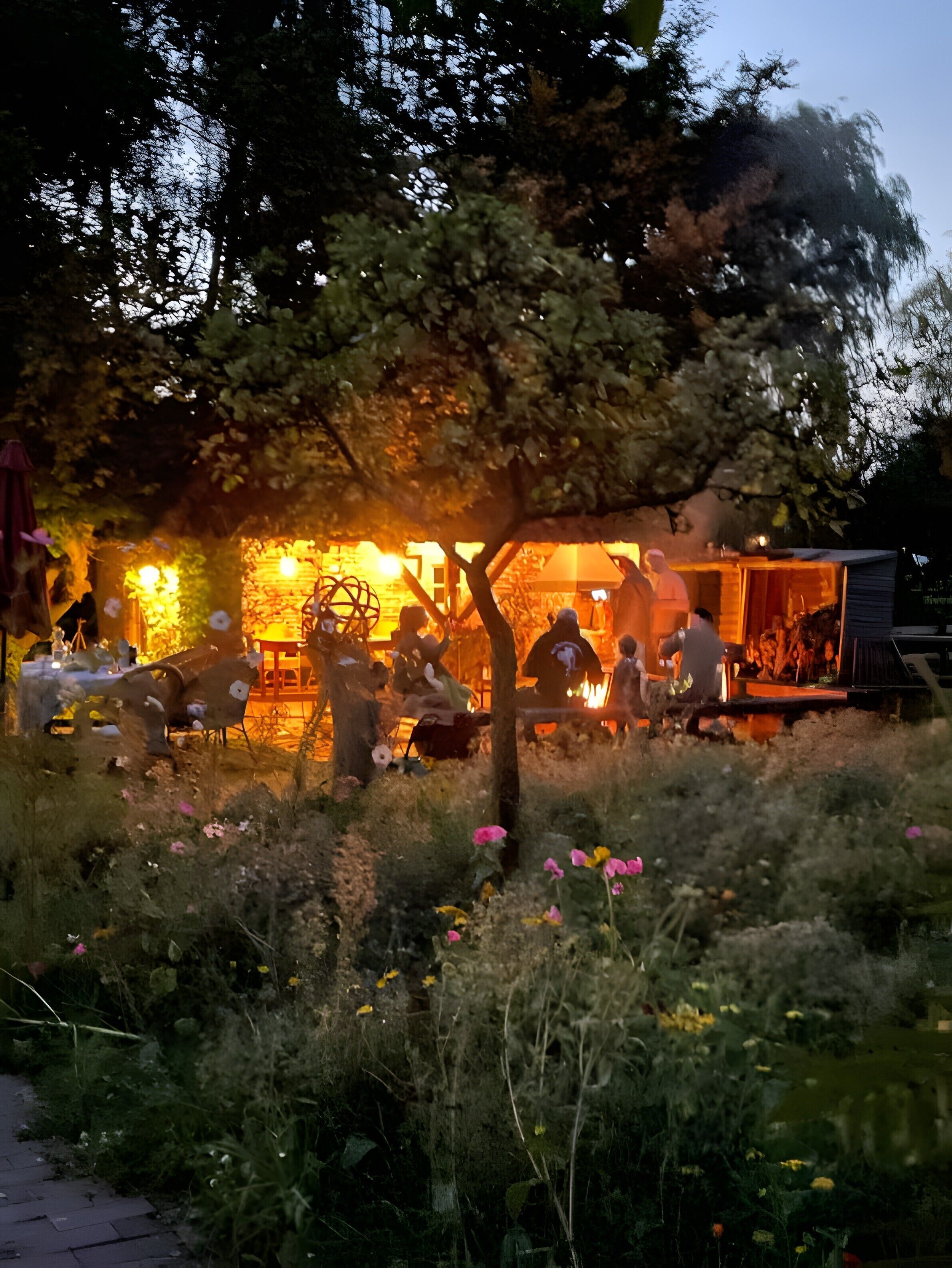 En uteservering med bord och stolar, omgiven av grönska och blommor.