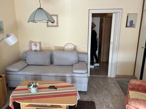 Ein Wohnzimmer mit einer Couch, einem Tisch mit gestreiftem Tischläufer, einer Lampe und einer Tür zu einem anderen Raum.