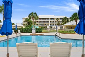 A swimming pool with lounge chairs and umbrellas, a poolside sign, and a multi-story building with balconies in the background.