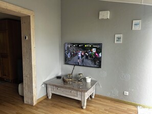 A room with a television mounted on the wall displaying a cycling event, a wooden table with decorative items, and a door leading to another room. - Small house with air conditioning and a small garden (Frontignan)