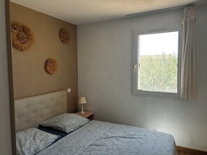 A bedroom with a bed, a nightstand, a lamp, and a window with a view of trees. - Small house with air conditioning and a small garden (Frontignan)