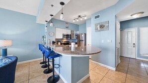 A modern kitchen with a central island, bar stools, and a dining area with a blue sofa. - High Tide Hideaway at Shores of Panama #1918 - Oceanfront, High-Rise, Pools, Covered Parking (Panama City Beach)