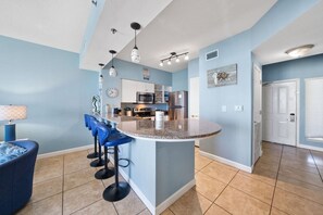 A modern kitchen with a central island, bar stools, and a dining area with a blue sofa. - High Tide Hideaway at Shores of Panama #1918 - Oceanfront, High-Rise, Pools, Covered Parking (Panama City Beach)