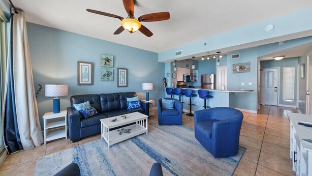 A living room with a sofa, coffee table, and chairs, a ceiling fan, and a kitchen area with bar stools. - High Tide Hideaway at Shores of Panama #1918 - Oceanfront, High-Rise, Pools, Covered Parking (Panama City Beach)