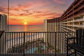 Een hoog hotel met uitzicht op de ondergaande zon boven de oceaan, een zwembad en een strand met parasols.