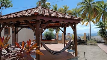A wooden gazebo with a tiled roof, surrounded by tropical plants and palm trees, overlooking a coastal area.