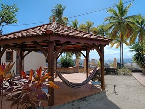 Un quiosco de madera con techo de tejas, rodeado de plantas tropicales y palmeras, con vista a la costa.