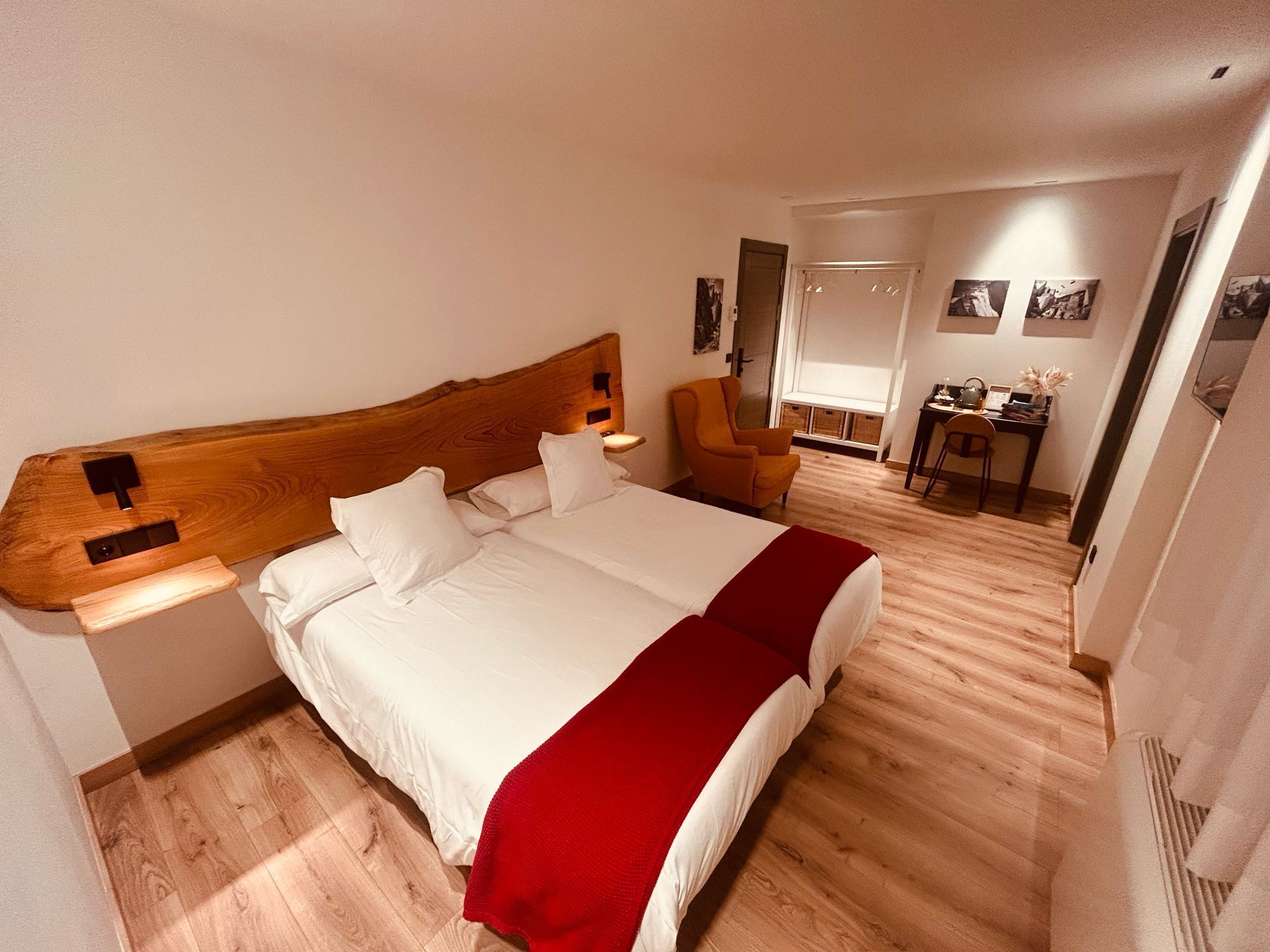 Habitación de hotel con una cama grande, cabecera de madera, manta roja, suelo de madera, un escritorio con silla y una ventana con persianas.
