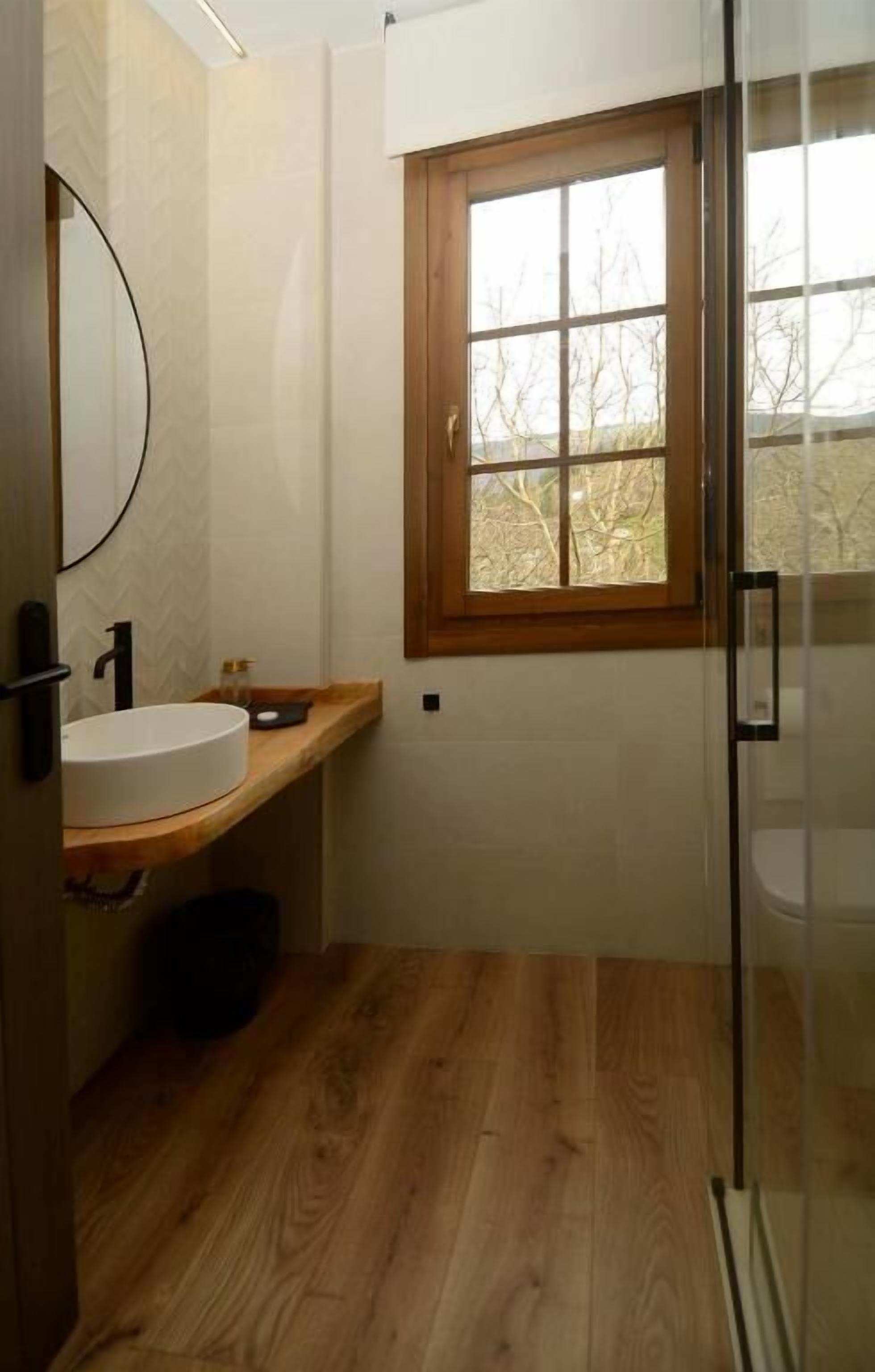 Un bany amb sòl de fusta, un lavabo rodó, una finestra i una mampara de vidre per a la dutxa.