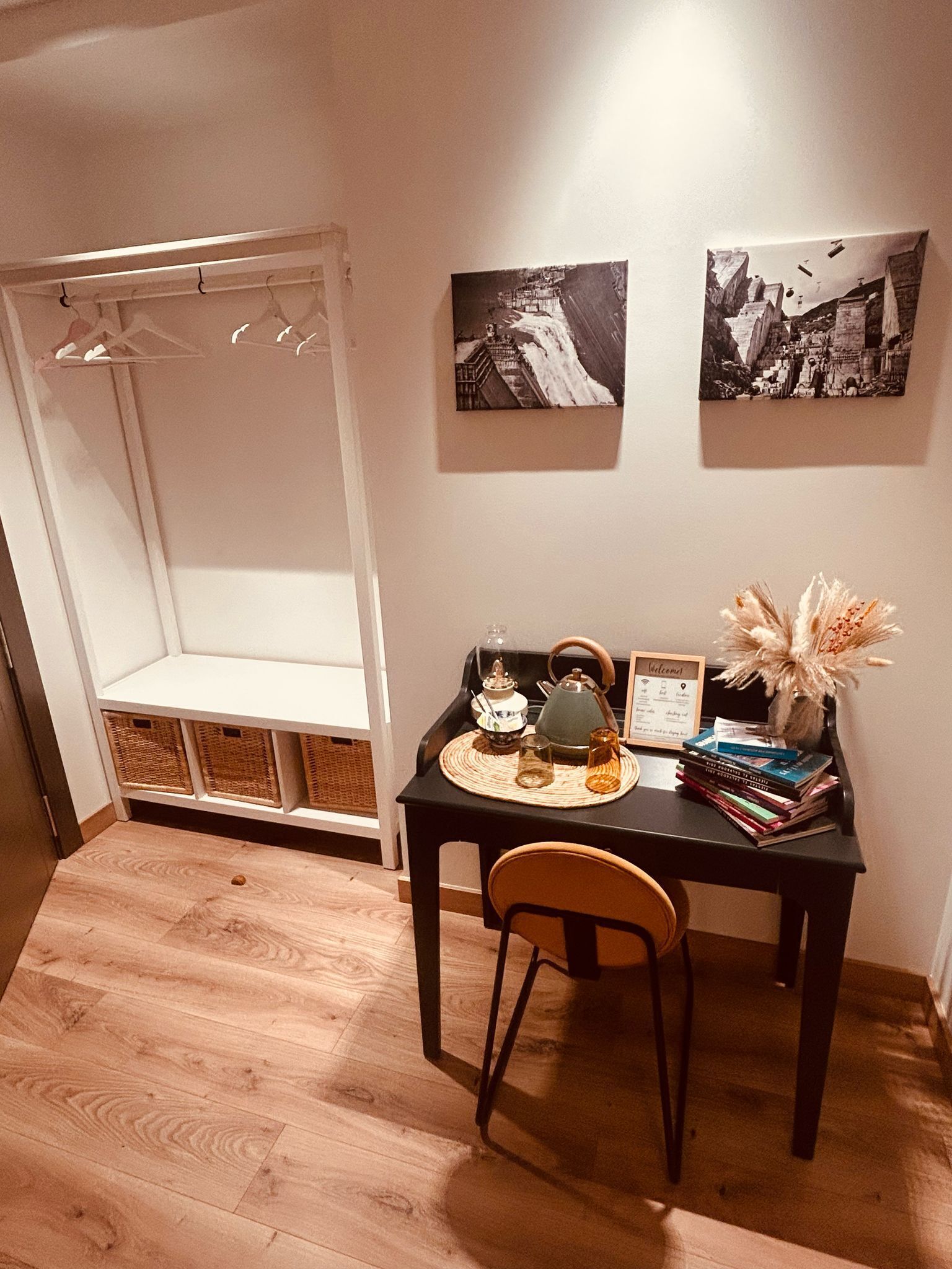 Una habitación con piso de madera, una mesa negra con una silla, un juego de té, un cartel de bienvenida y dos cuadros en la pared.