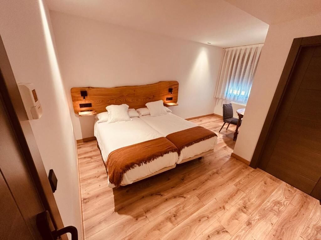 Habitación de hotel con cabecera de madera, cama con ropa blanca, mesita de noche y una silla.